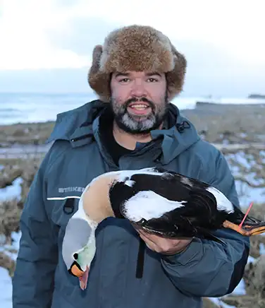 A hunter in camouflage kneeling outdoors holding a shotgun next to a turkey.